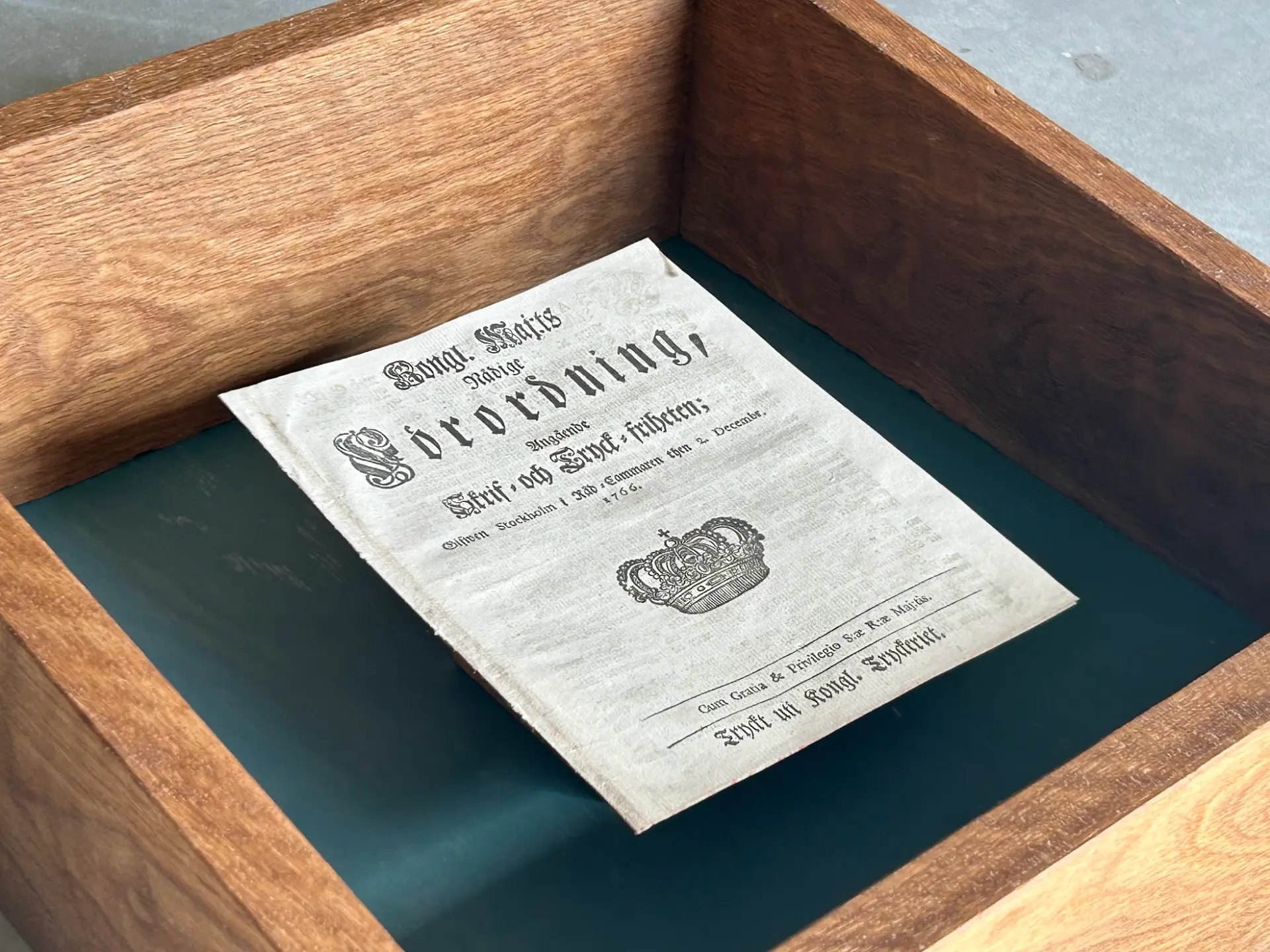 1766 års tryckfrihetsförordning liggande i en öppen trämonter med mörkt blågrön botten.
